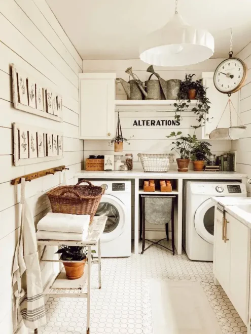 A cozy and well-organized laundry room with white walls and appliances. There are wooden shelves with labeled storage bins, framed pictures on the wall, a vintage scale, and potted plants adding a touch of greenery. A wicker basket sits on a folding ironing board beside the washing machine.