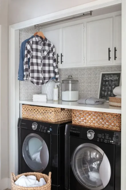 An organized laundry room with stacked black washer and dryer units, a white countertop with jars, towels, and wicker baskets, white cabinets above, and a plaid shirt hanging on a hook.