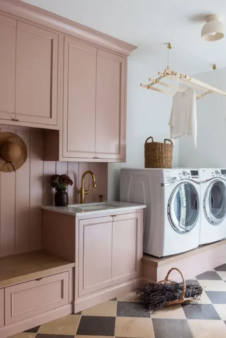 Laundry room with soft pink cabinetry, black and white checkered floor, side-by-side washer and dryer, wooden hanging rack, and open shelving with decorative items and plants.