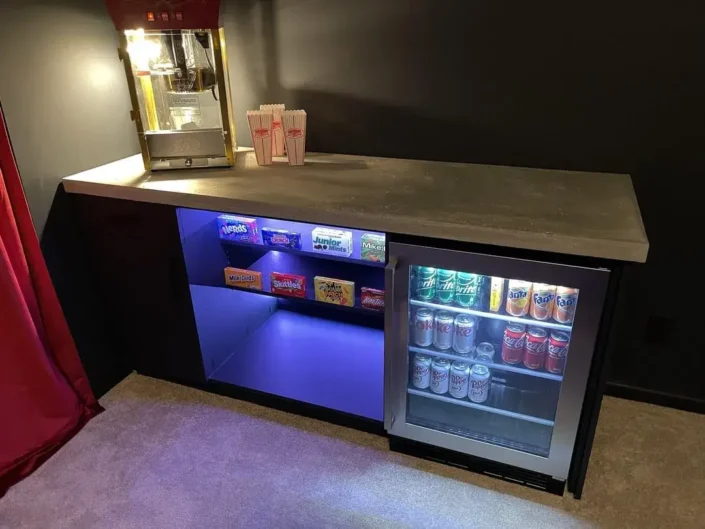 Home theater snack bar with a dark countertop, blue under-counter lighting, and a selection of candy displayed behind glass panels, including M&M’s, Skittles, and Reese’s, along with a glass-front refrigerator stocked with canned beverages and a popcorn machine, perfect for enhancing a basement home theater room with authentic cinema-style concessions