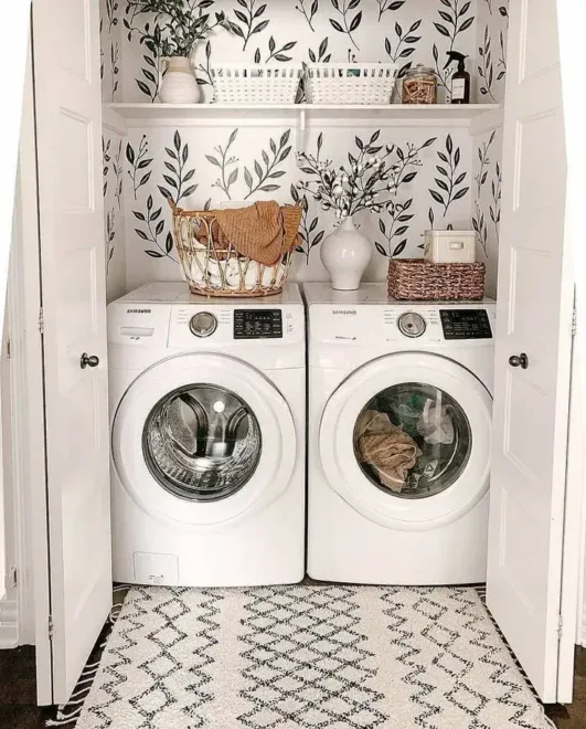 Stacked front-loading washing machines with a shelf above holding decorative items and plants, set against leafy vine wallpaper.